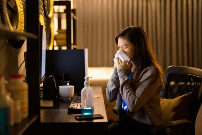 Sick young Asian woman blowing nose with tissue while working from home late at night